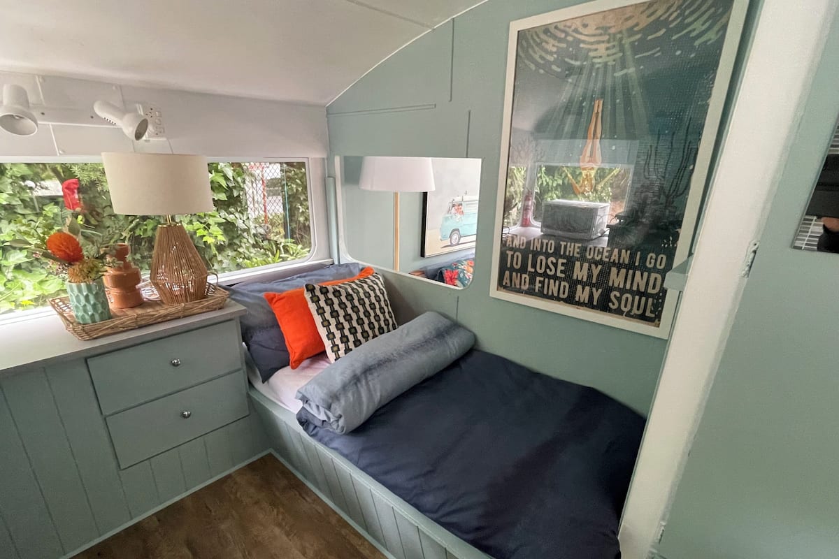 A compact bedroom with a bed, a window, a small table with decorative items, and a poster on the wall.