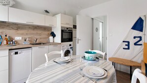 A modern kitchen with white cabinets, a dishwasher, an oven, and a sink. There is a dining table set with plates, glasses, and a bowl. - Studio "Port Lazo" with Sea View, Private Terrace and Wi-Fi (Plouézec)