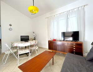 Un soggiorno con un divano, un tavolino in legno, una TV su un mobile in legno, un piccolo tavolo da pranzo con sedie e una lampada a sospensione gialla.