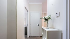 En hall med en vit dörr längst in, ett vitt konsolbord med en vas med blommor och en väggmonterad termostat.
