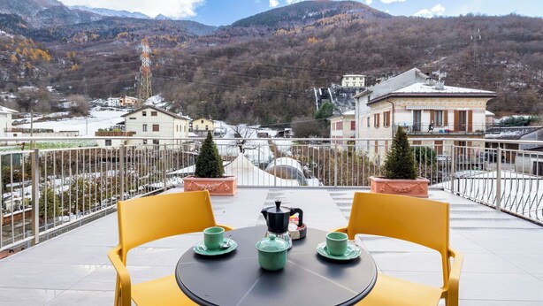 Una terraza con sillas amarillas y una mesa, con vistas a un pueblo y montañas.