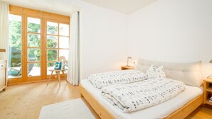 Ein Schlafzimmer mit einem hölzernen Bett, einem Sessel, einem Nachttisch und Blick durch eine Glastür auf einen Außenbereich.