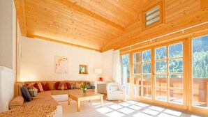 Ein gemütliches Wohnzimmer mit Holzbalkendecke, einer Couch mit bunten Kissen, einem Couchtisch und einem großen Fenster mit Blick ins Freie.