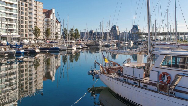 Un port de plaisance où sont amarrés de nombreux bateaux, un grand immeuble doté de balcons et un ciel d’un bleu limpide.