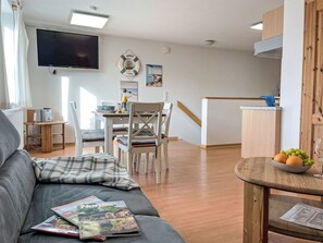 Ein Wohnzimmer mit grauem Sofa, ein Essbereich mit Tisch und Stühlen, eine Küche mit Holzschränken und einem an der Wand montierten Fernseher.