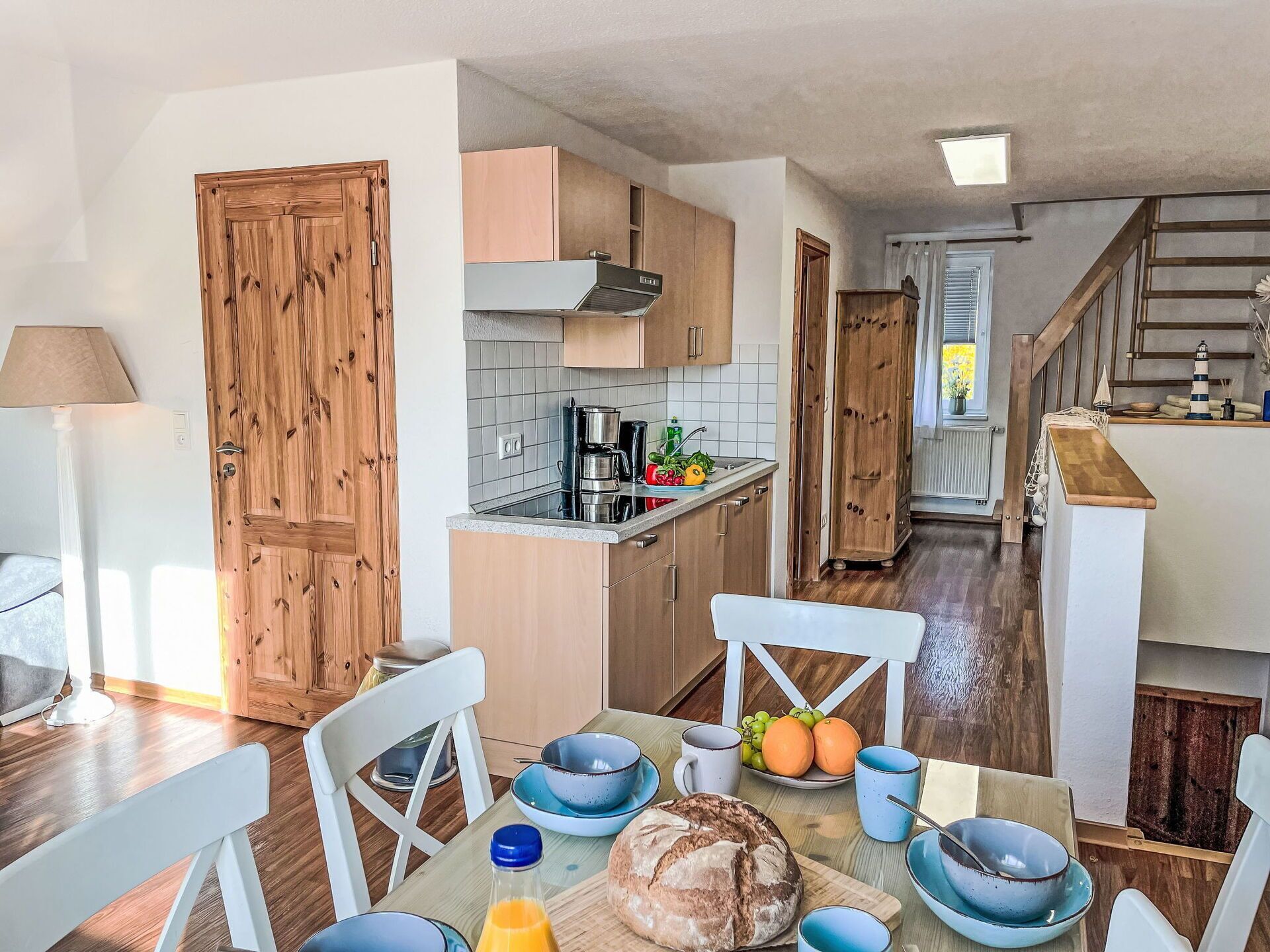 Cocina compacta con armarios de madera, comedor con mesa dispuesta para dos personas y una escalera que sube a otro nivel.