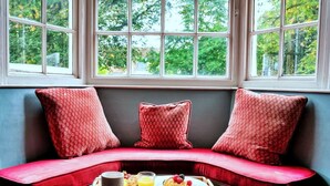 A cozy seating area with a red cushioned bench, a tray with breakfast items, and a view of greenery through large windows.