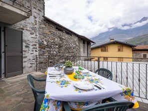 Un espace extérieur aménagé pour les repas, avec un mur en pierre, un bâtiment jaune et une vue sur la montagne.