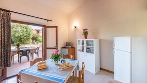 Un coin repas avec une table dressée pour deux personnes, un buffet blanc aux portes vitrées, un réfrigérateur blanc et une porte-fenêtre coulissante donnant sur une terrasse extérieure.