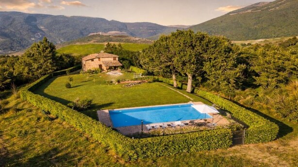 A pool surrounded by a well-maintained lawn, a stone building, and trees in a hilly landscape. - VILLA NASCA - Ville Carborello (Castelraimondo)