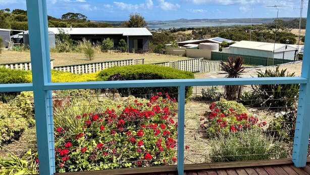 Vista de uma varanda com grade de metal, com vista para um jardim com flores vermelhas e uma paisagem distante com edifícios e um corpo de água.