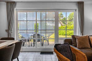 A room with a dining table, chairs, a sofa, and a large sliding glass door leading to an outdoor area with a patio and greenery.