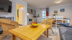 Un soggiorno moderno con un tavolo da pranzo in legno, sedie in vimini, un divano grigio, una TV a schermo piatto e una cucina sullo sfondo.