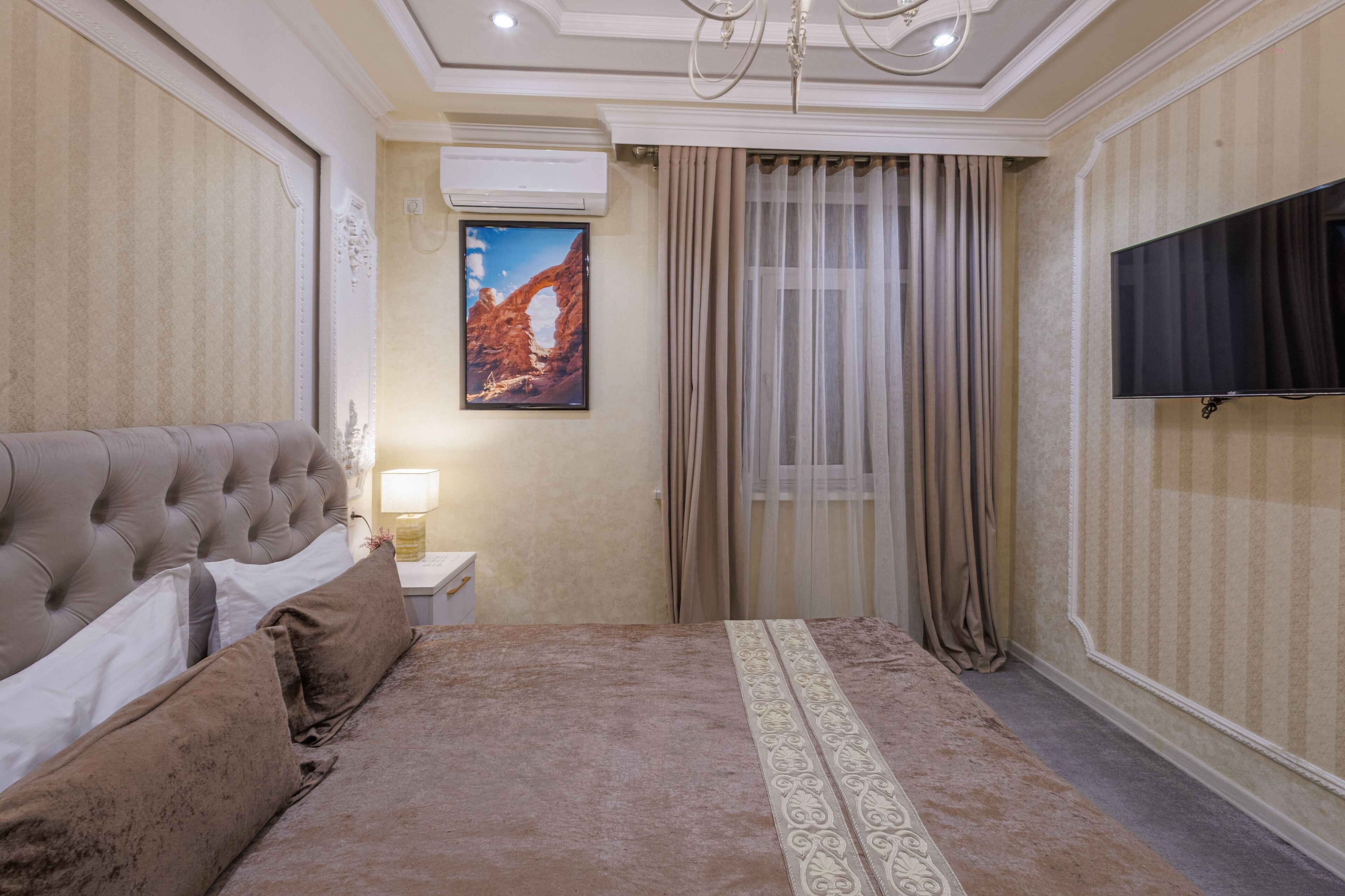 A bedroom with a tufted headboard, a flat-screen TV, a painting of a canyon, and a window with curtains.