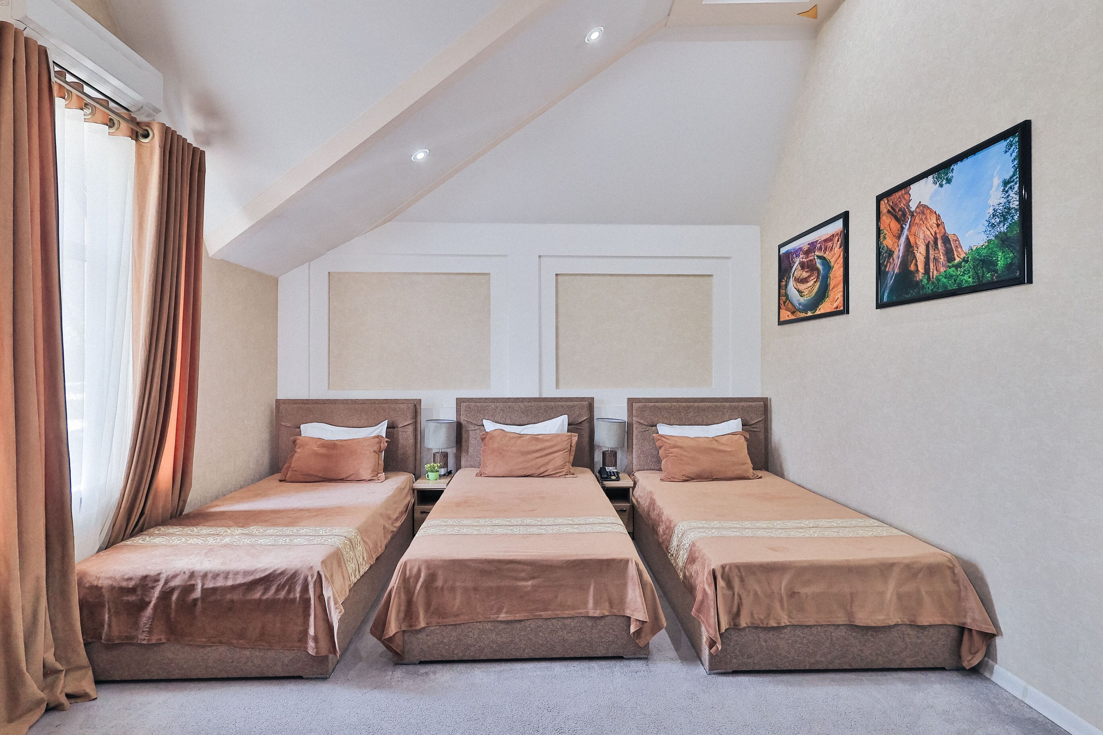 A hotel room with three single beds, a large window with curtains, and two framed pictures on the wall.