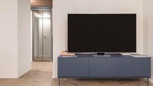 Ett modernt vardagsrum med en stor platt-TV som är monterad på ett grått skåp, ett trägolv och en hall som leder till ett annat rum.
