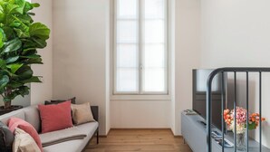 Un salon moderne avec un canapé gris, une grande plante verte, une fenêtre et un meuble pour la télévision agrémenté d’un vase de fleurs.