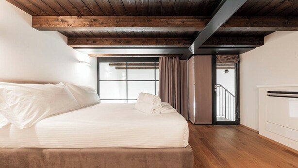 Une chambre à coucher avec un lit, des poutres en bois au plafond, une fenêtre et une porte donnant sur un escalier.