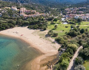 Vista aérea de uma área costeira com praia, águas cristalinas e área residencial com edifícios e vegetação.