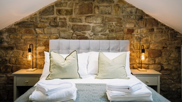 A bedroom with a stone wall, a tufted headboard, two bedside tables with lamps, and neatly folded towels. - Sun St Apartment 2 (Lancaster)