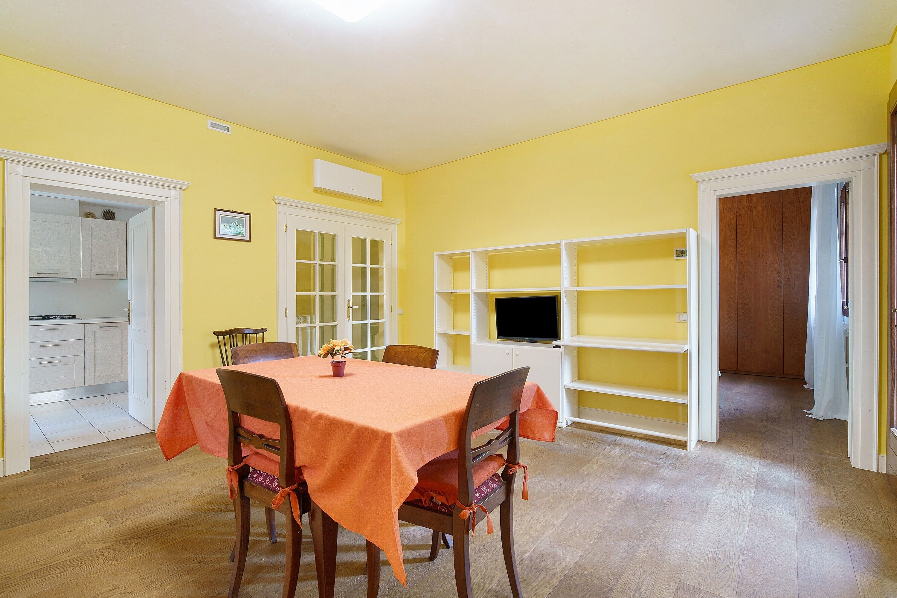 Un comedor con una mesa dispuesta para cuatro personas, rodeada de sillas, una estantería y una cocina visible a través de un umbral abierto.