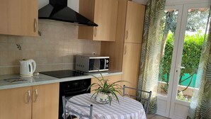 A compact kitchen with wooden cabinets, a gas stove, a microwave, and a kettle. There is a small table with a checkered tablecloth and two chairs. A potted plant is on the table.