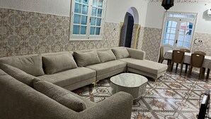Una sala de estar con un sofá seccional color crema, una mesa de centro redonda, baldosas de suelo con diseño, una zona de comedor con mesa y sillas, y un techo decorativo.