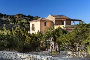 Une maison en pierre à deux étages, avec un toit en tuiles, entourée d’arbres et d’un mur en pierre.