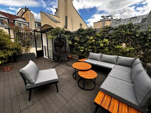 Ein Sitzbereich im Freien mit einer Couch, Stühlen und Tischen, umgeben von einem Garten und einem Gebäude.
