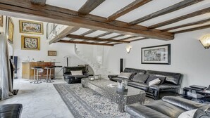 Un soggiorno ampio con soffitto alto, travi in legno a vista, un divano in pelle nera, un tappeto decorato e una scala che conduce a un piano superiore.