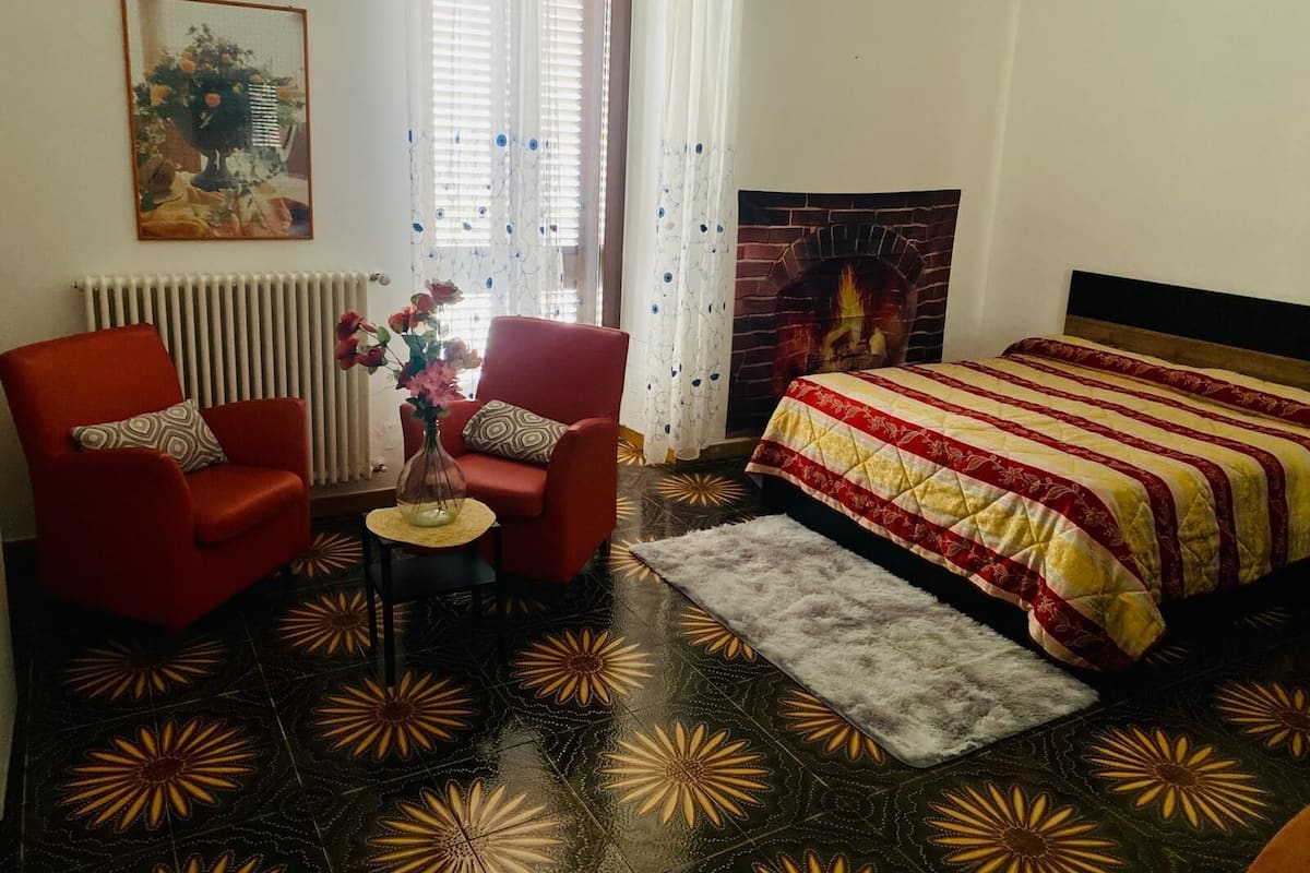 Una stanza con un letto, due poltrone rosse, un tavolino con un vaso, un termosifone, un caminetto e un pavimento decorato.