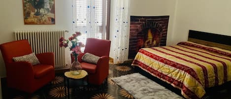 A room with a bed, two red chairs, a small table with a vase, a radiator, a fireplace, and a patterned floor.
