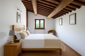 Ein Schlafzimmer mit Holzboden, einem Bett mit weißer Bettdecke, einem Nachttisch mit Lampe und einem Fenster mit Blick ins Grüne.
