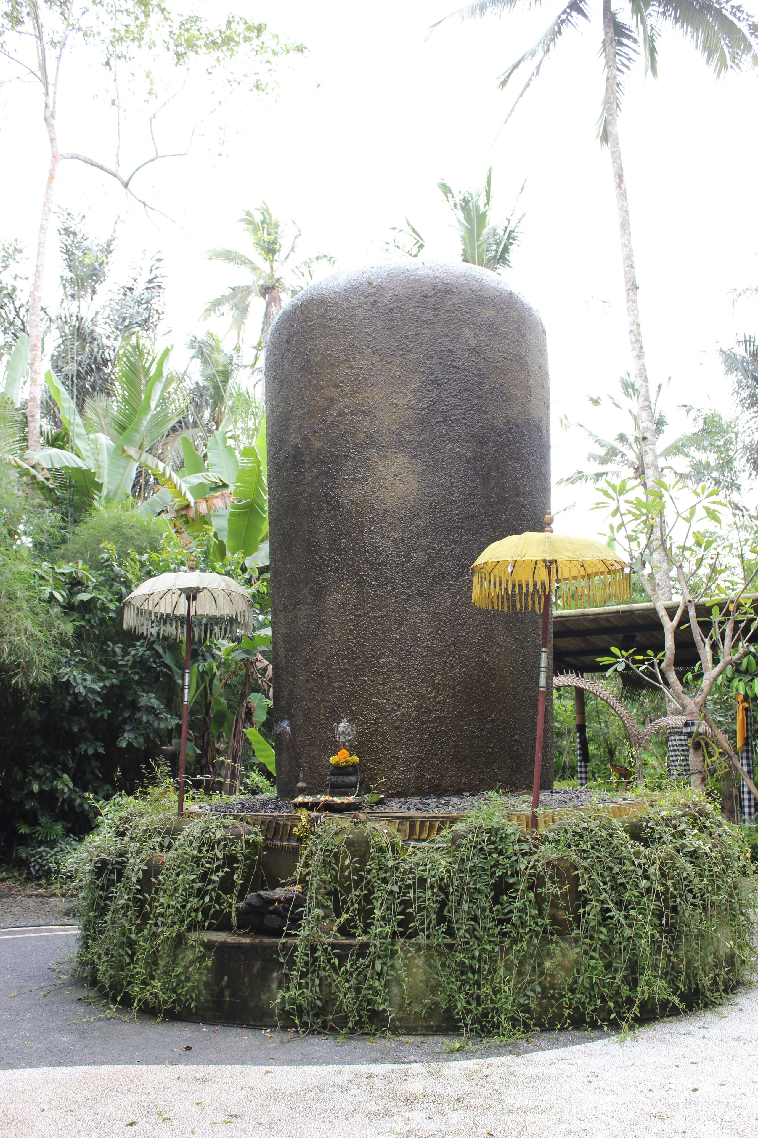 Une grande structure cylindrique en pierre, entourée d’une végétation luxuriante et agrémentée de deux parasols.