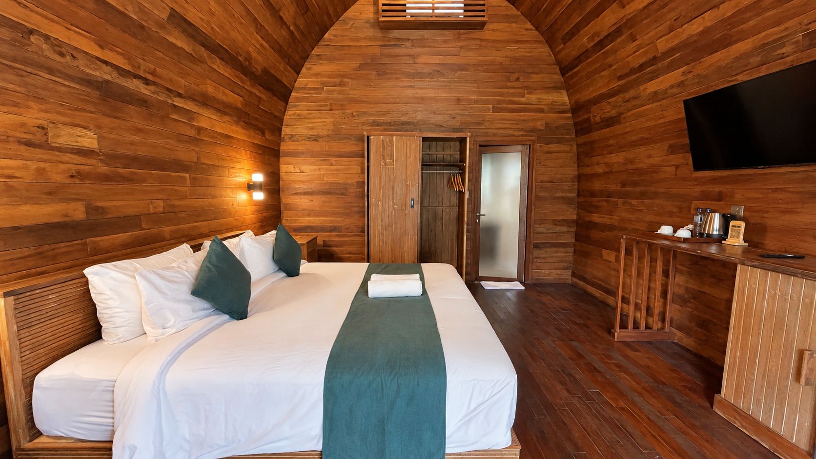 Una habitación con techo de madera, una cama con ropa de cama blanca y almohadas verdes, un televisor de pantalla plana montado en la pared y un mueble de madera con varios objetos encima.