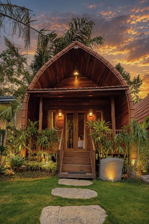 A wooden cabin with a porch, surrounded by tropical plants and a stone pathway leading to the entrance. - The Jungle Pots (Payangan)