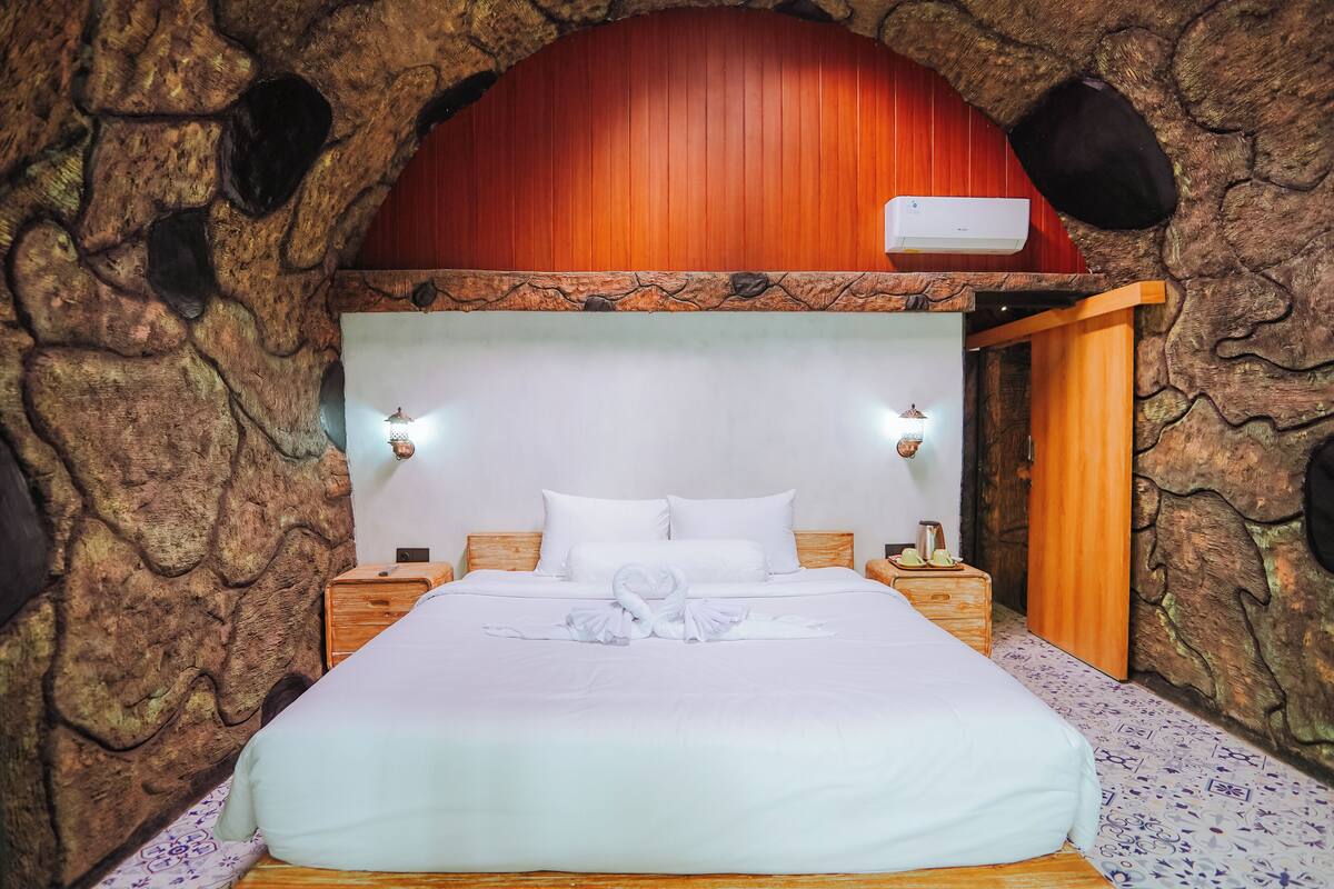 A bed with white linens and a towel art arrangement, wooden headboard, two bedside tables, and a stone wall backdrop.