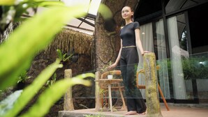 A woman standing near a glass railing, with a stone wall and a wooden chair in the background. - The Jungle Pots (Payangan)