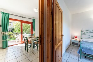 A room with a wooden door, a bed, a small bedside table, and a view of a balcony with a table and chairs. - Cozy Accommodation in Beautiful Sardinia (Badesi)