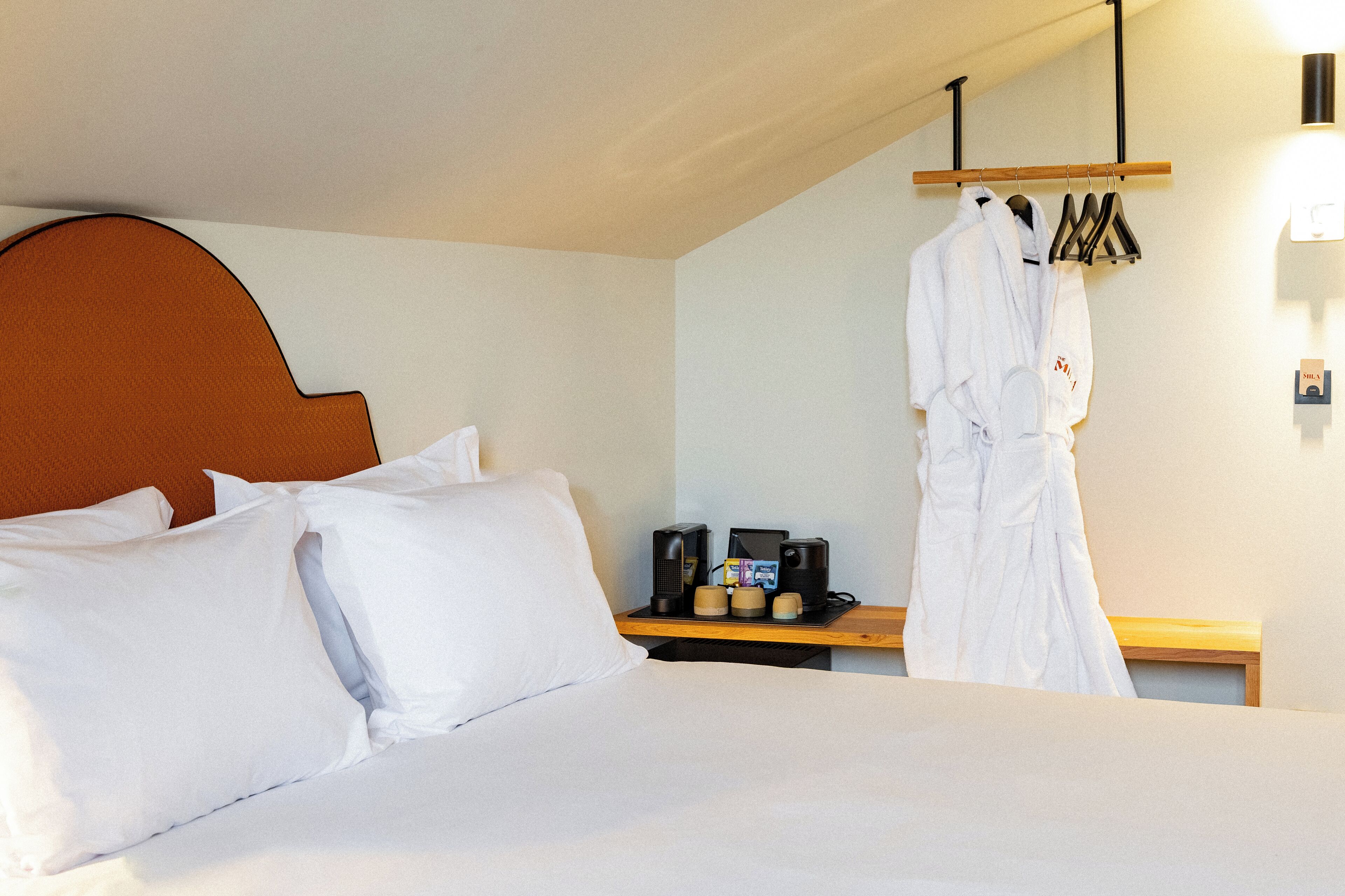 Une chambre d’hôtel comprenant un lit, des oreillers, une table de chevet avec une machine à café, un peignoir et un porte-manteau.