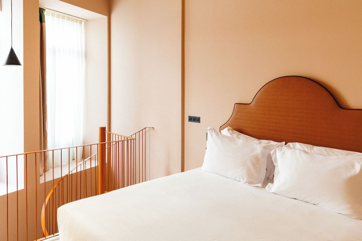 A hotel room with a bed, a window with curtains, a railing, and a wall switch.