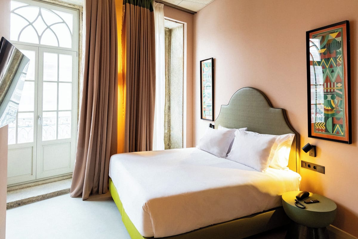 A hotel room with a bed, a nightstand, a window with curtains, and a wall-mounted artwork.