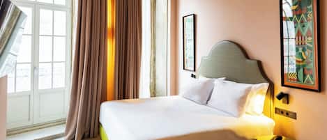 A hotel room with a bed, a nightstand, a window with curtains, and a wall-mounted artwork.