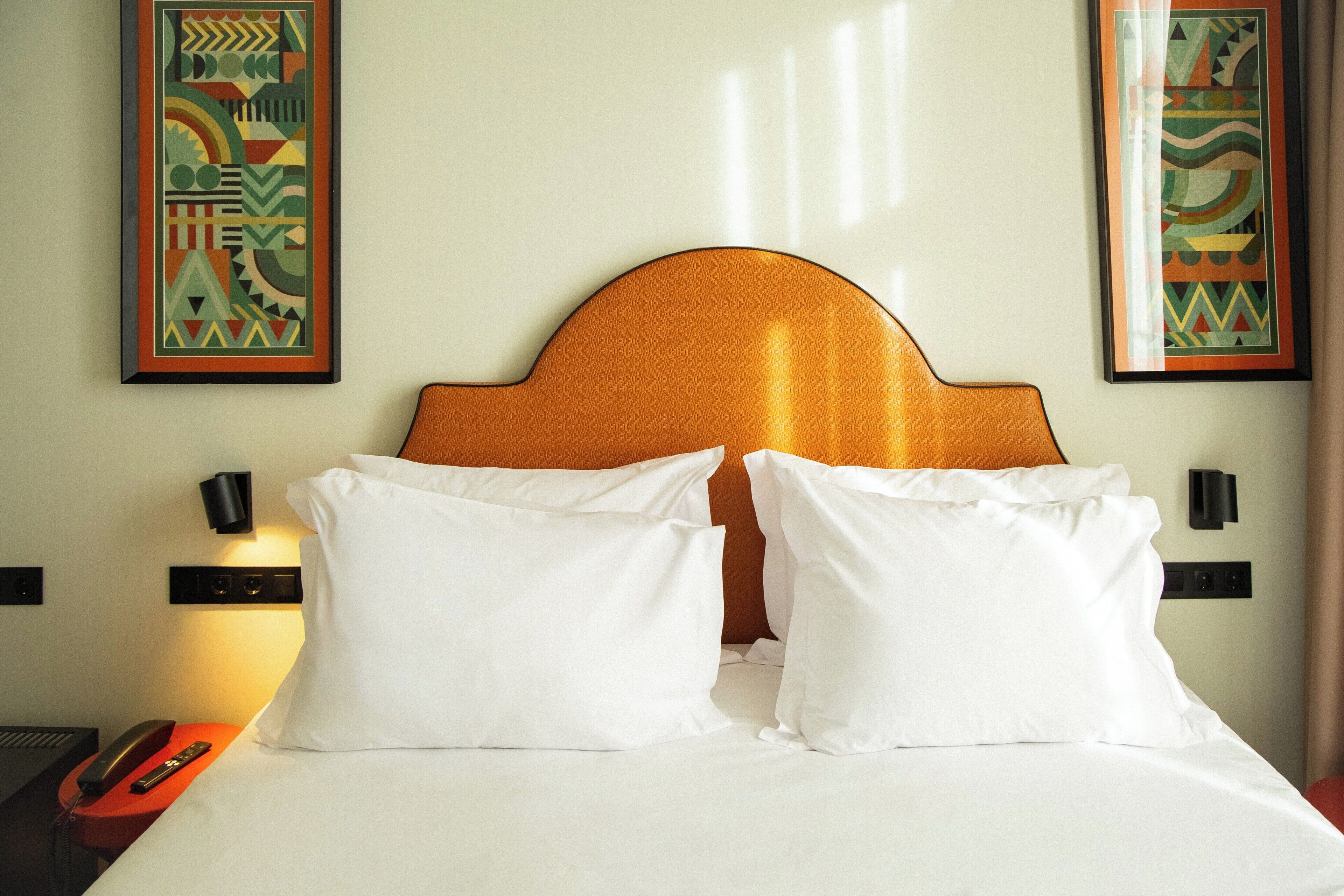 A hotel room with a bed, two pillows, a nightstand with a phone and remote, and two wall-mounted lamps.
