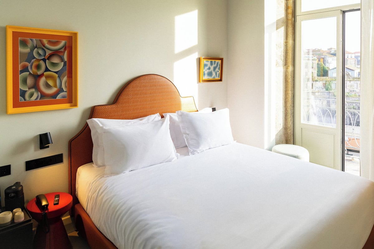 A hotel room with a bed, a nightstand, a red side table, a lamp, and two framed artworks on the wall.