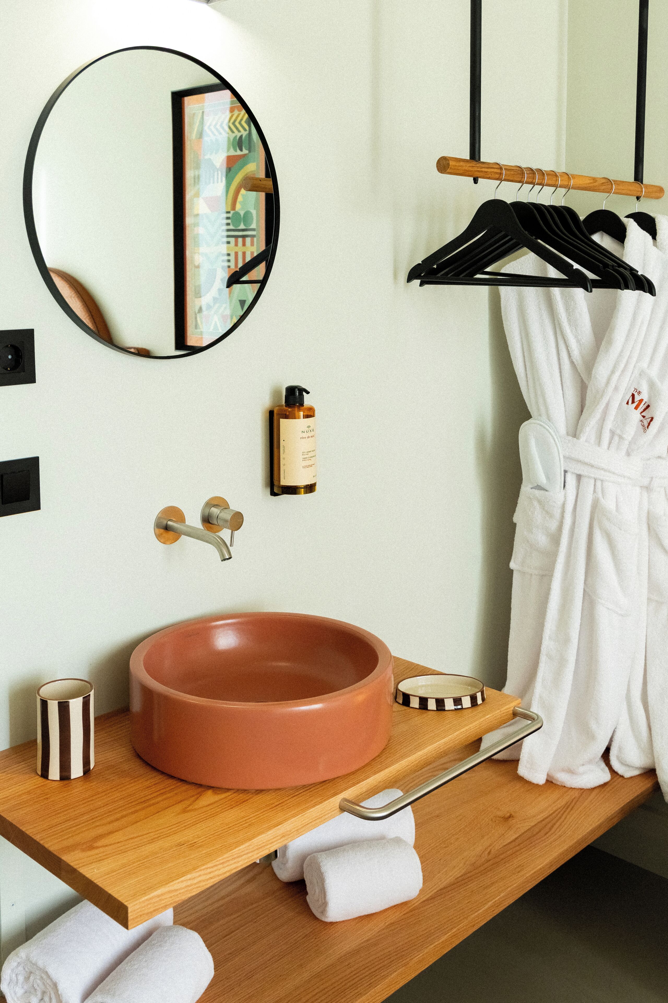 A bathroom with a round sink, a mirror, a towel rack, and a robe hanging.