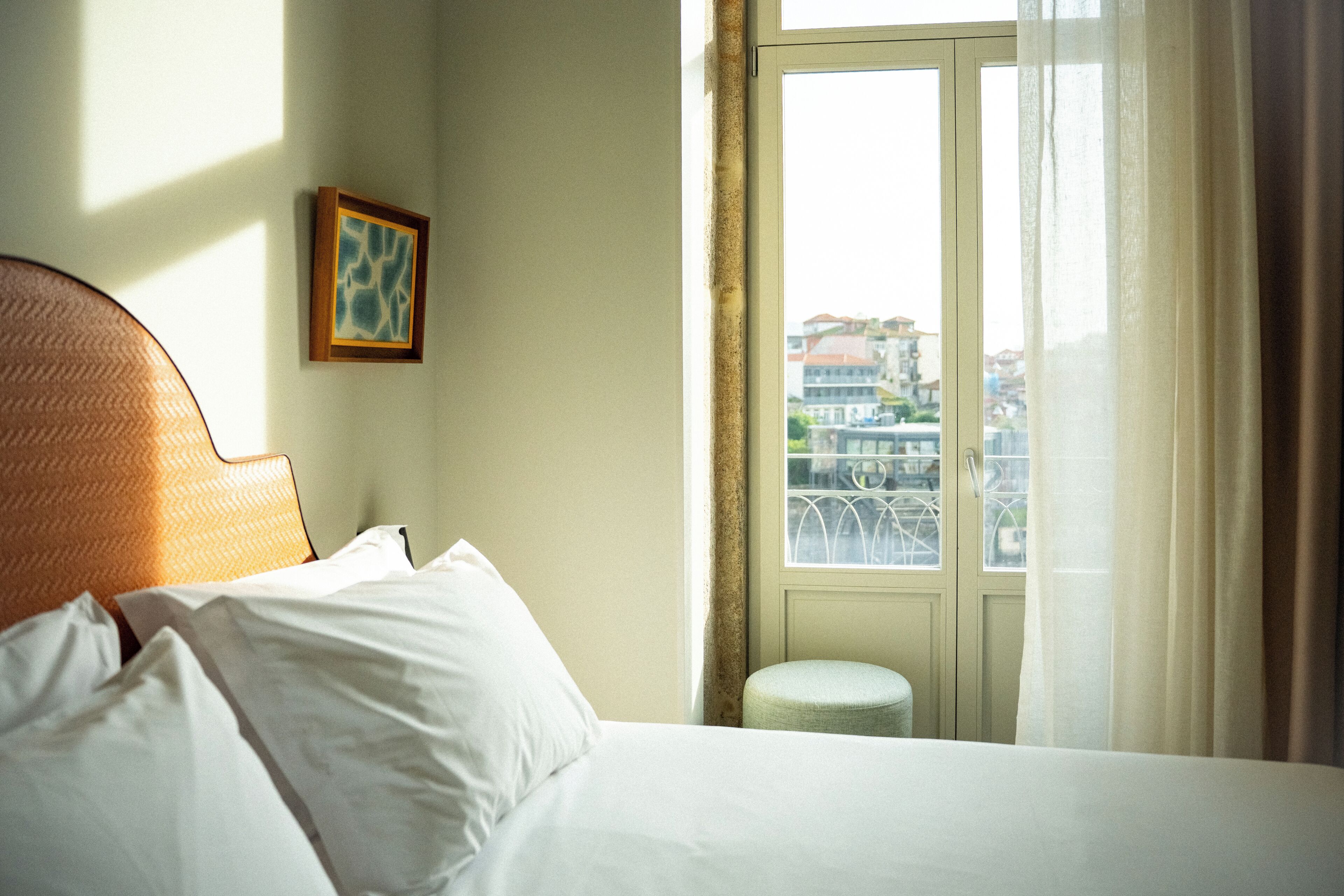 A hotel room with a bed, a small stool, a window with a view of buildings, and a framed picture on the wall.
