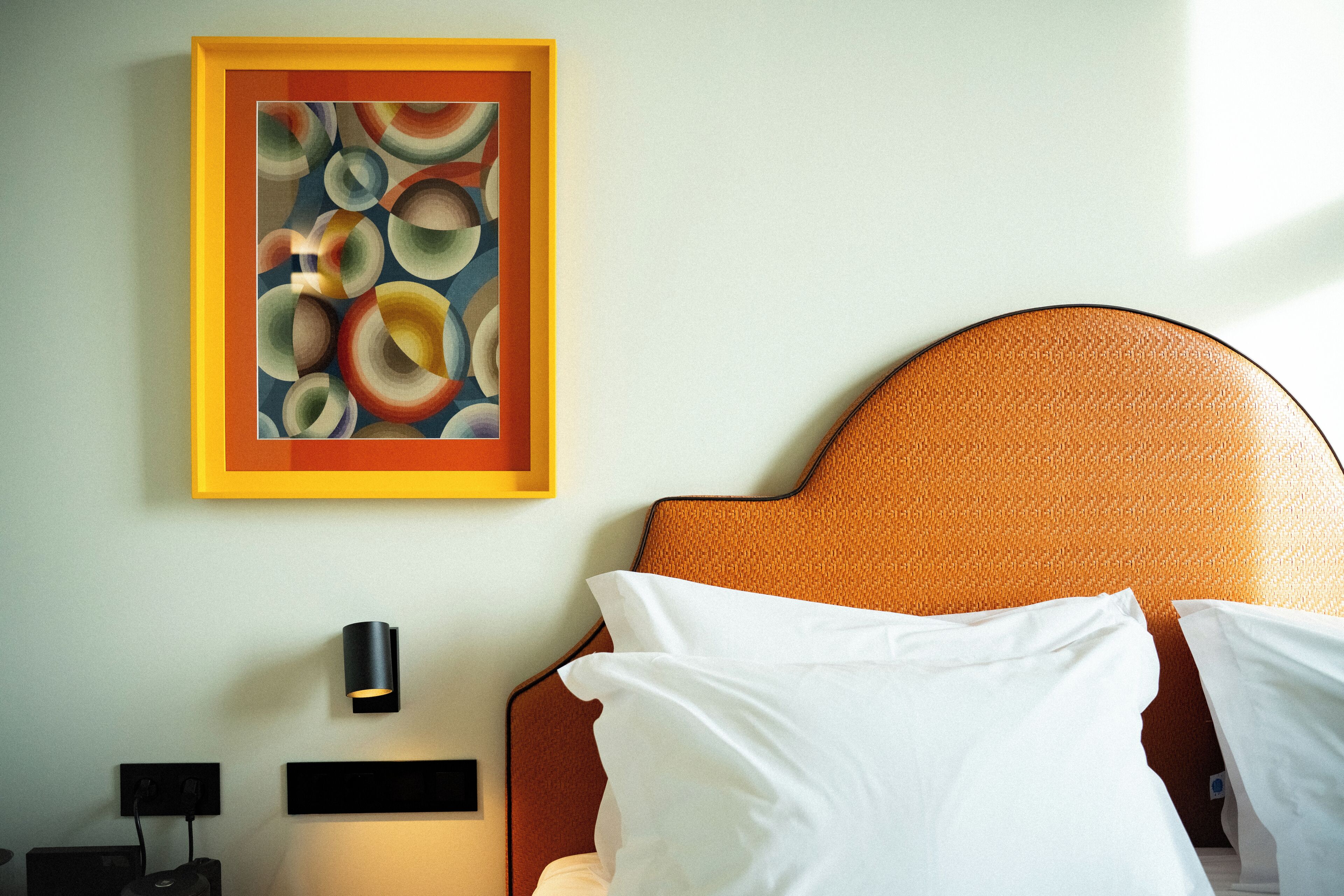 A hotel room with a bed, a painting on the wall, a nightstand, and a wall-mounted lamp.