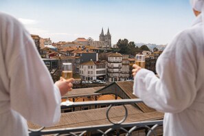 Dos personas sostienen copas en un balcón con vistas a un paisaje urbano que incluye edificios históricos y una catedral destacada.