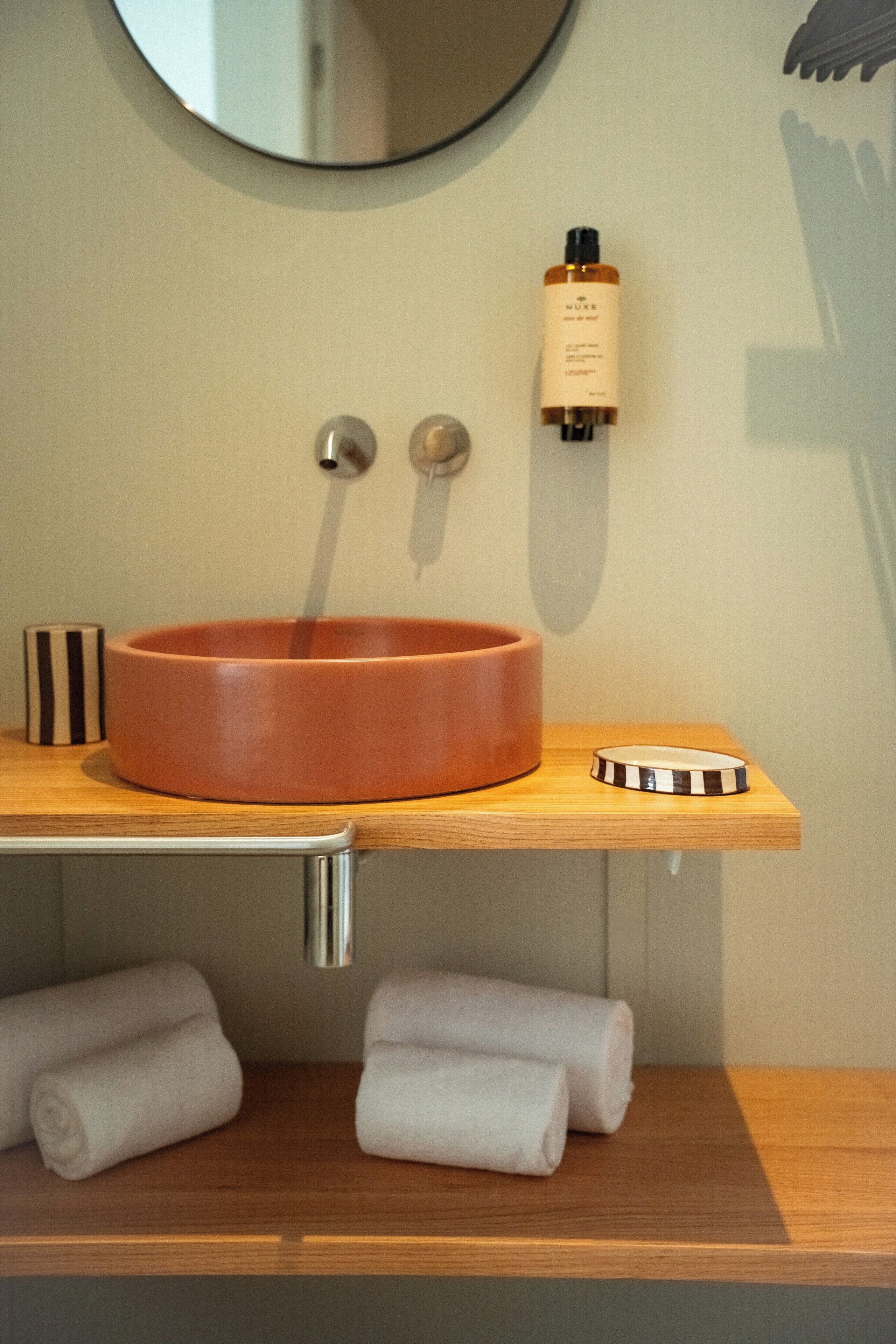 A bathroom with a round ceramic sink, a mirror above it, a soap dispenser, and two rolled towels on a wooden shelf.
